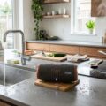 Vandtæt Bluetooth højttaler placeret på køkkenbord ved opvaskekummen, designet til fugt og varme